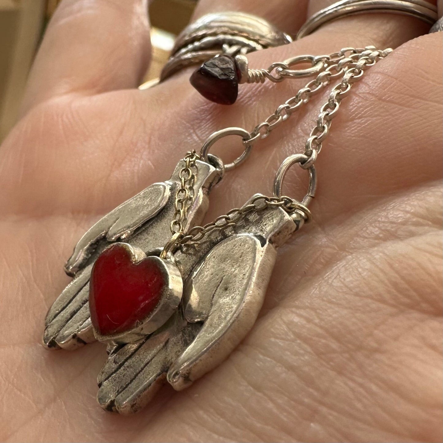A gorgeously simple pair of fine silver hands cupping a red enamelled heart attached by 9ct gold chain hung on a sterling silver chain.