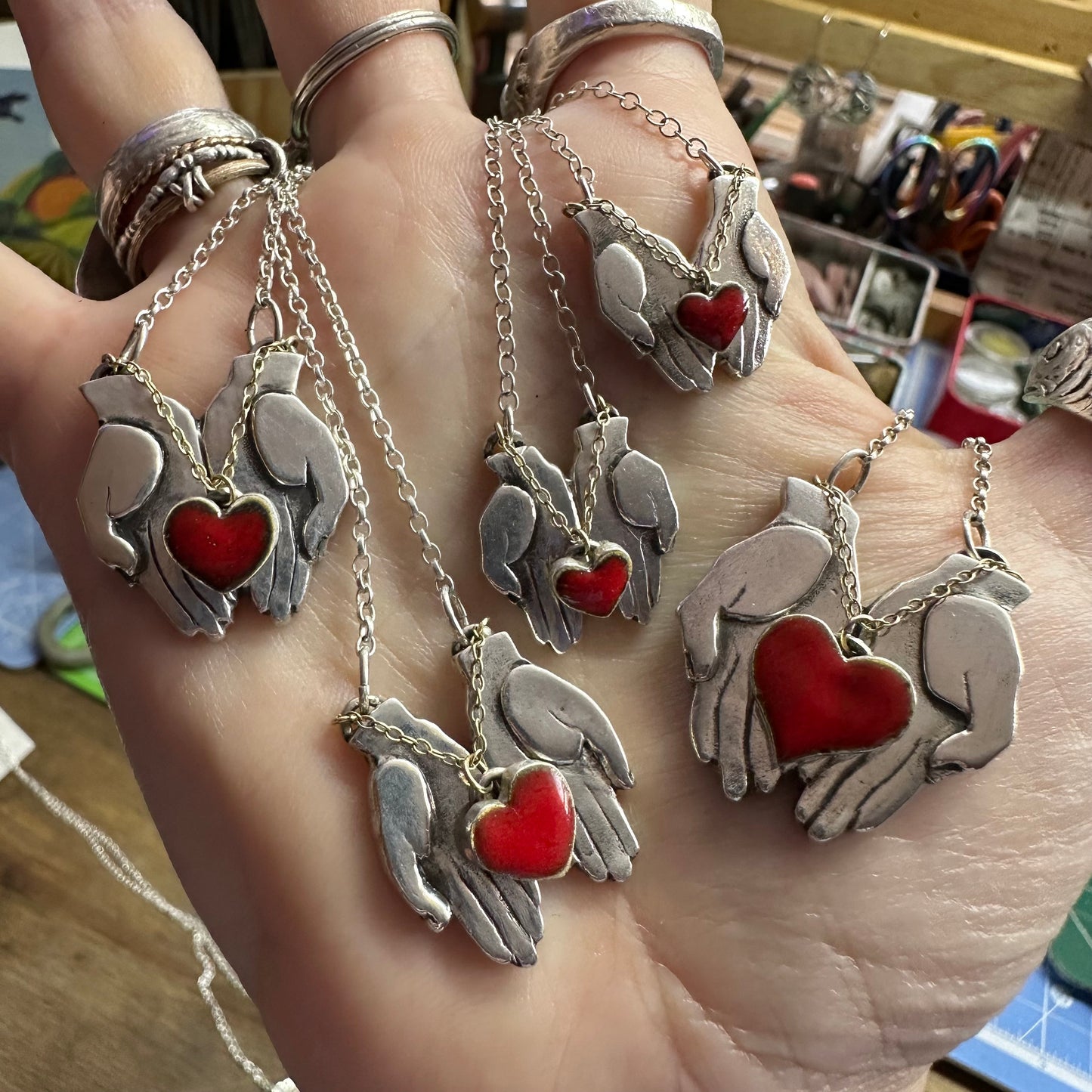 A gorgeously simple pair of fine silver hands cupping a red enamelled heart attached by 9ct gold chain hung on a sterling silver chain.