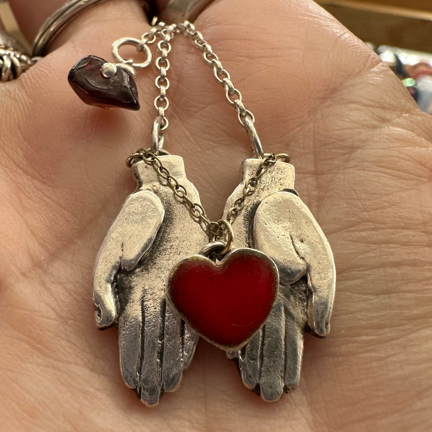 A gorgeously simple pair of fine silver hands cupping a red enamelled heart attached by 9ct gold chain hung on a sterling silver chain.