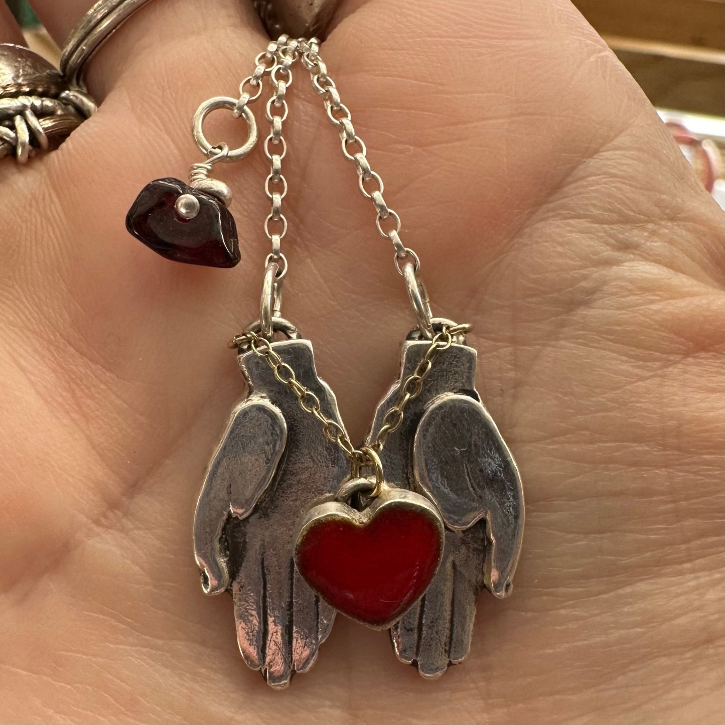 A gorgeously simple pair of fine silver hands cupping a red enamelled heart attached by 9ct gold chain hung on a sterling silver chain.