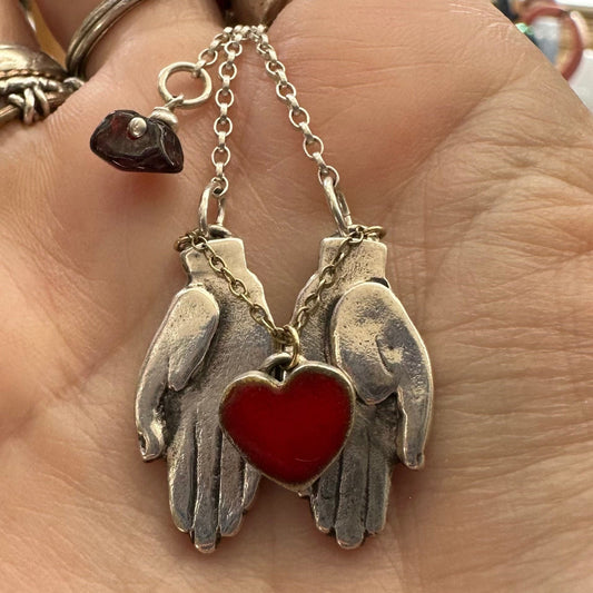 A gorgeously simple pair of fine silver hands cupping a red enamelled heart attached by 9ct gold chain hung on a sterling silver chain.